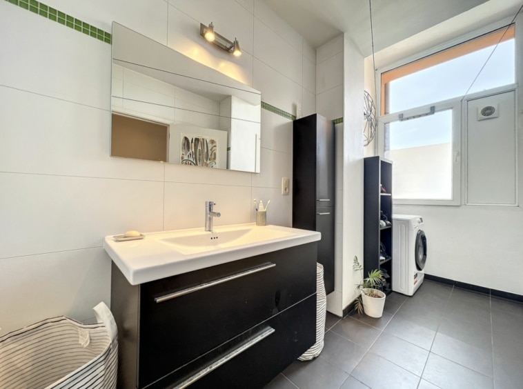Salle de bain moderne minimaliste avec éclairage naturel, vanity blanc, rangement noir et plante verte.
