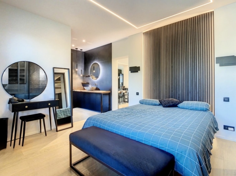 Chambre moderne et relaxante avec lit bleu, mur en bois, miroir rond, étagères lumineuses.