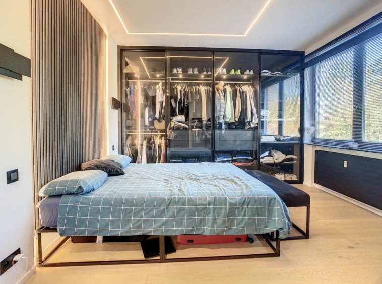 Chambre minimaliste lumineuse avec lit en métal, armoire vitrée et grandes fenêtres naturelles.