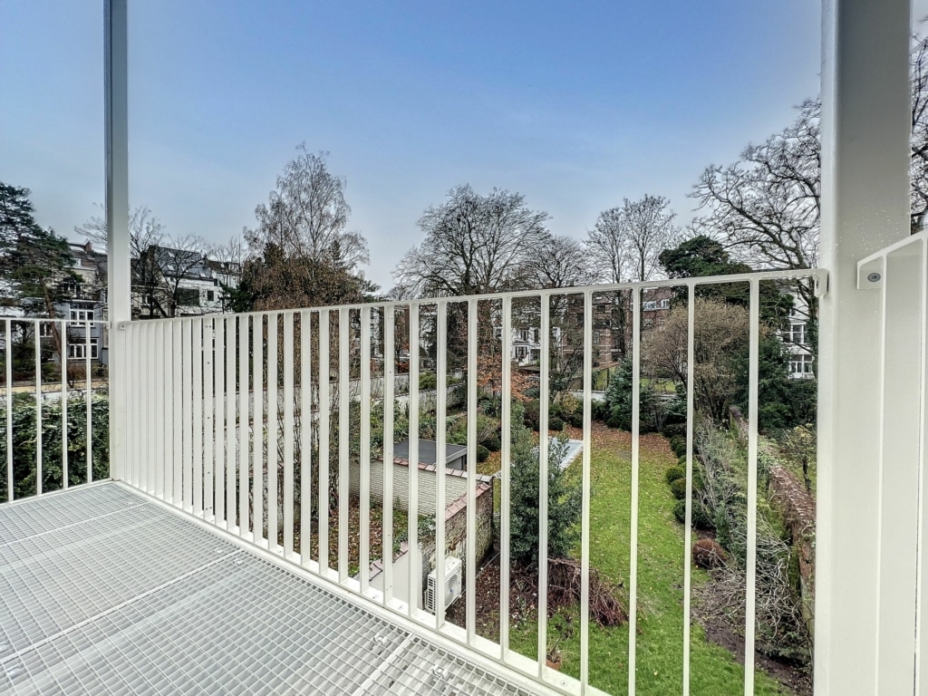 Balcon contemporain avec vue sur jardin verdoyant, pelouses, arbres et fleurs sous ciel bleu.