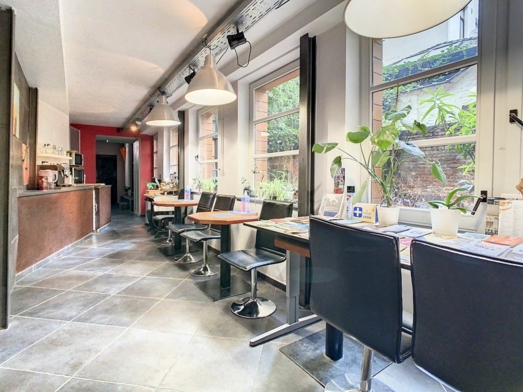 Café moderne lumineux avec tables en bois, plantes vertes et mur rouge, ambiance chaleureuse et relaxante.