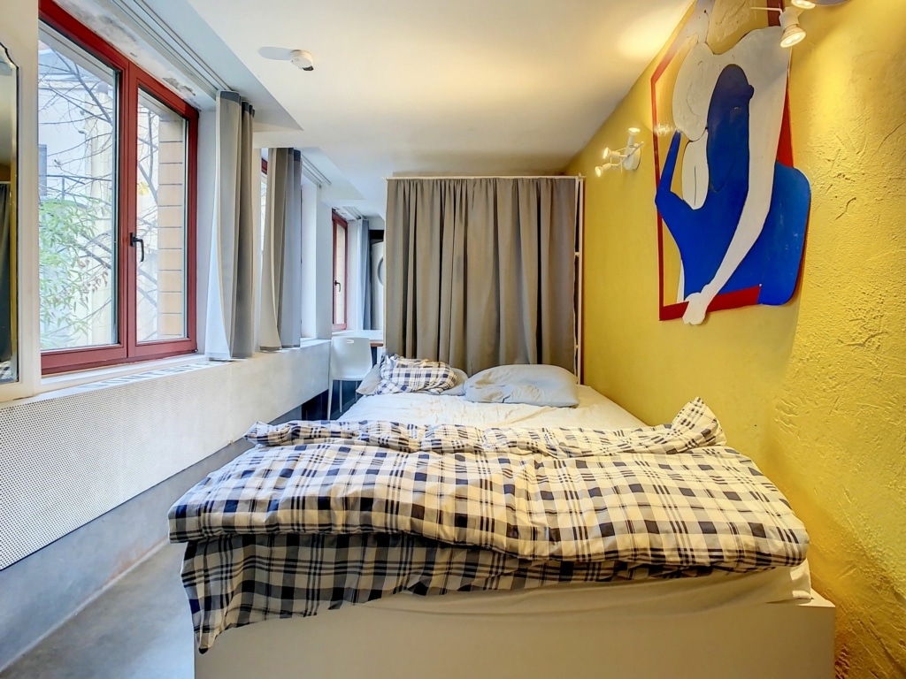 Chambre minimaliste et cosy avec murs jaunes, lit surélevé, fenêtres lumineuses et décoration colorée.