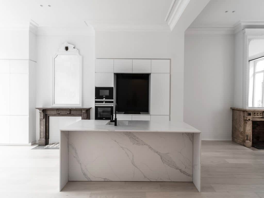Cuisine moderne lumineuse avec îlot en marbre, rangements blancs, cheminée en pierre et technologie intégrée.