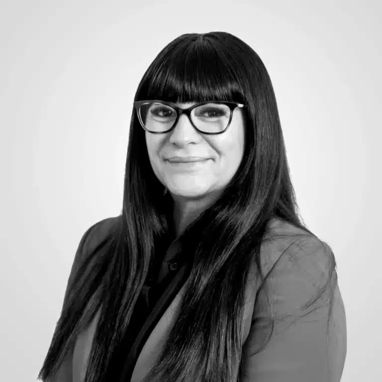 Femme professionnelle élégante portant lunettes modernes, blazer ajusté, sourire confiant, fond gris neutre.