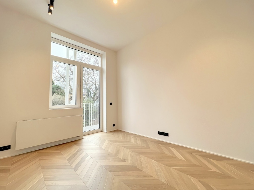 Espace lumineux minimaliste avec parquet en chevrons, grande fenêtre, vue verte et accès balcony.