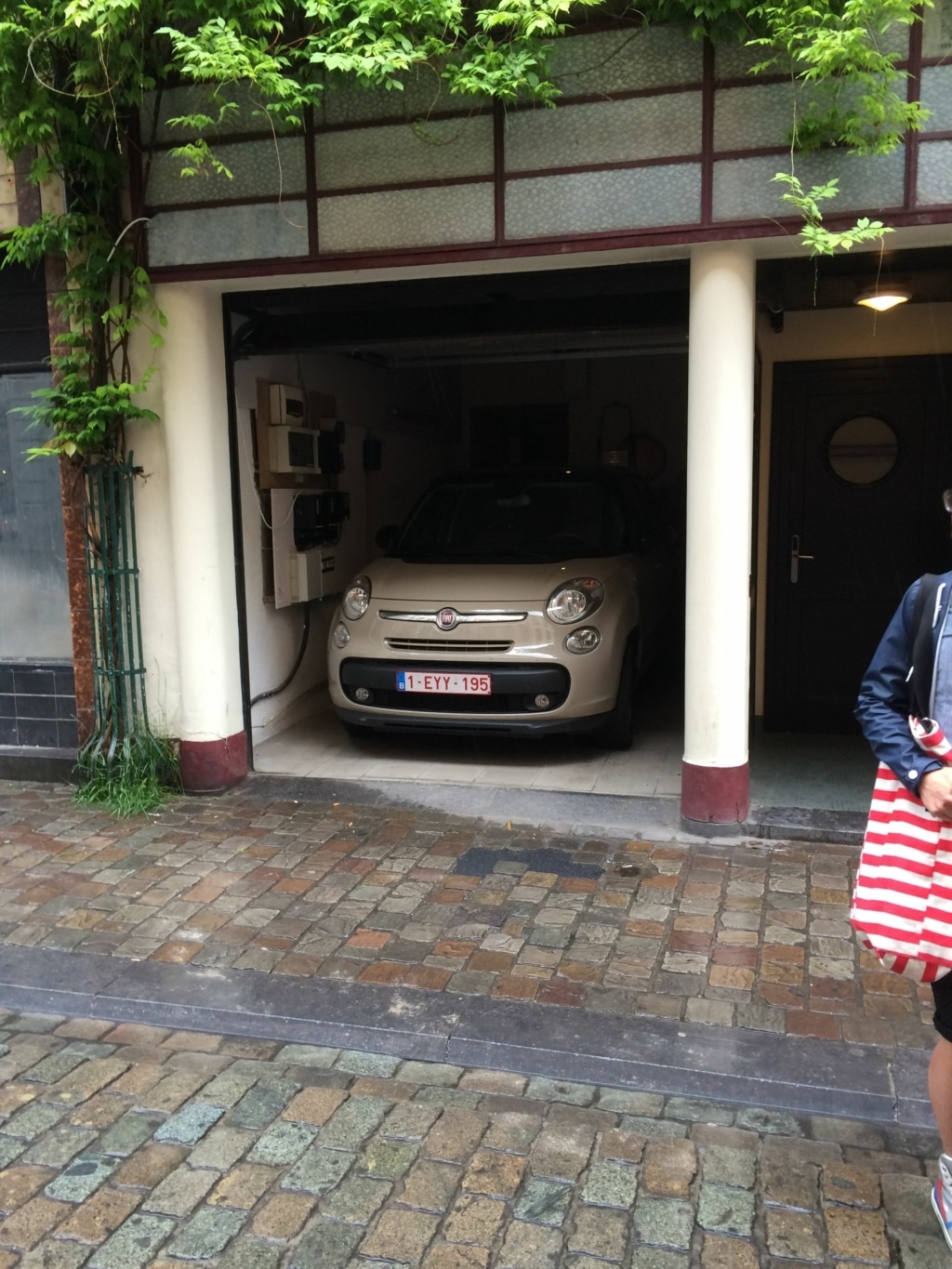 Rue pavée charmante avec garage traditionnel, Fiat vintage beige et végétation luxuriante.