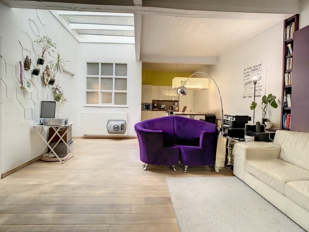 Salon lumineux avec mobilier moderne, espace de travail, plantes vertes et cuisine colorée.