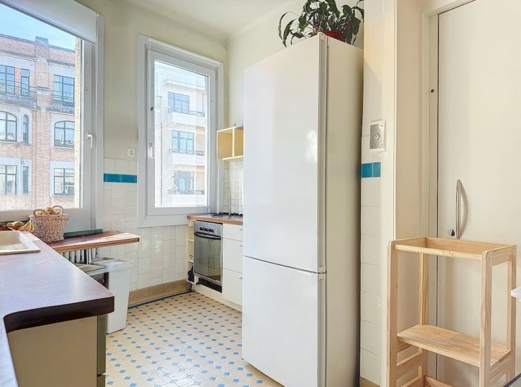 Cuisine moderne lumineuse avec réfrigérateur blanc, plan en bois, grandes fenêtres, style épuré.