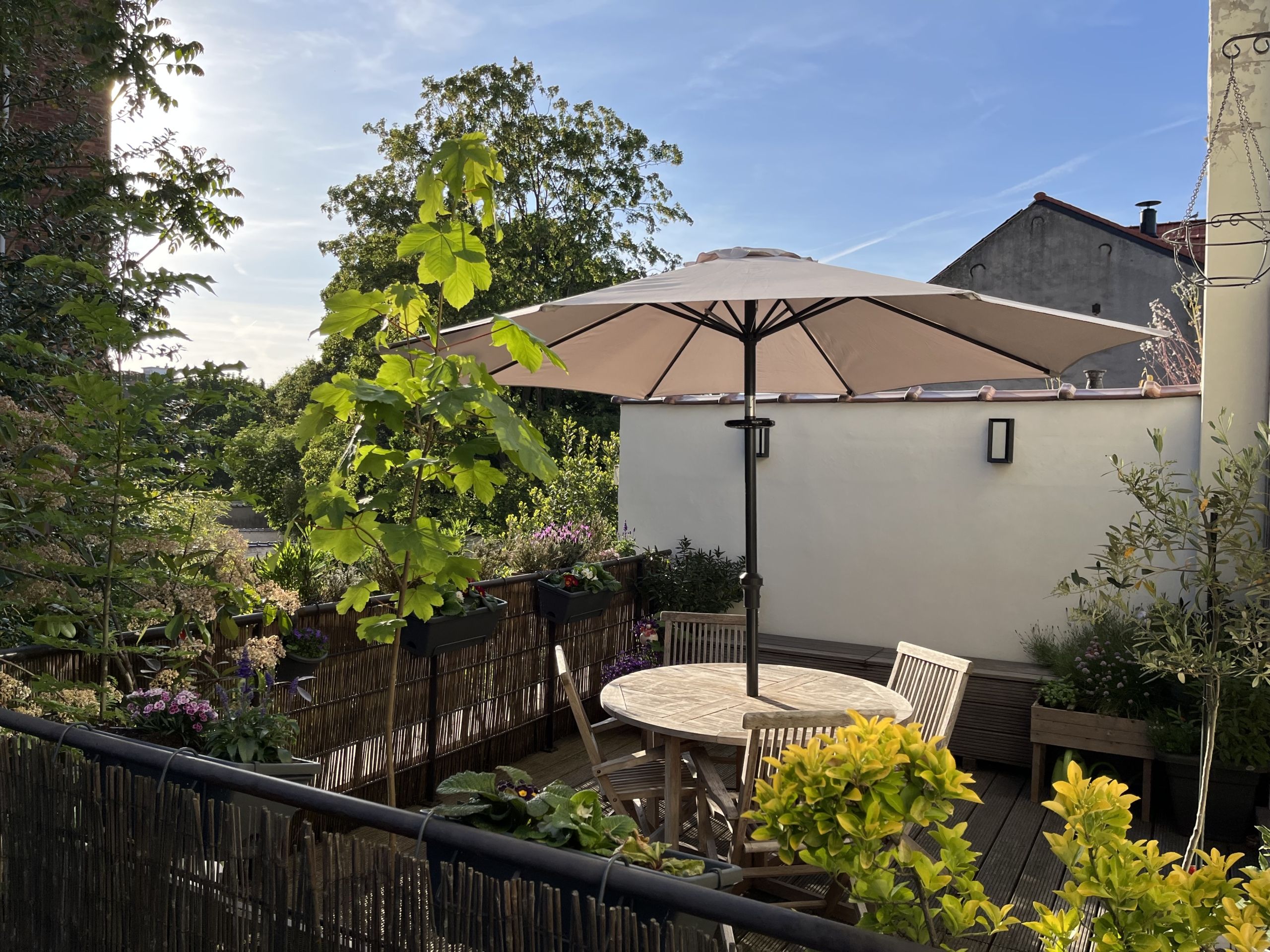 Espace extérieur cosy avec mobilier en bois, parasol beige, plantes vertes et fleurs colorées.