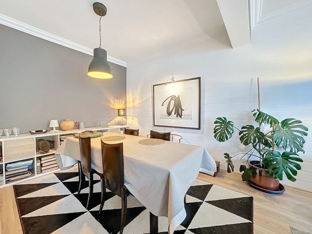 Salle à manger moderne avec table en bois clair, chaises foncées, art abstrait et végétation verte élégante.