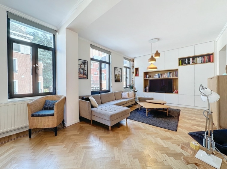 Salon lumineux avec canapé gris, chaise en osier, tapis foncé et étagères décoratives.