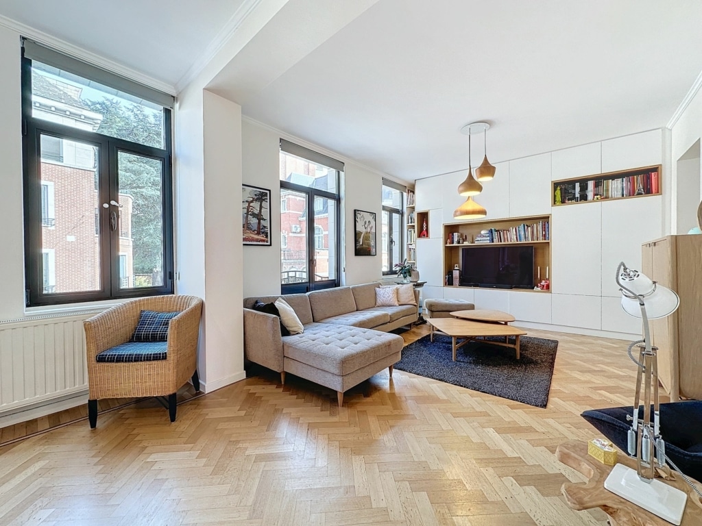 Salon lumineux avec canapé gris, chaise en osier, tapis foncé et étagères décoratives.