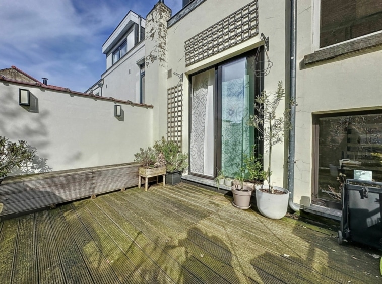 Terrasse urbaine moderne avec bois, grandes baies vitrées et plantes en pot, ambiance lumineuse.