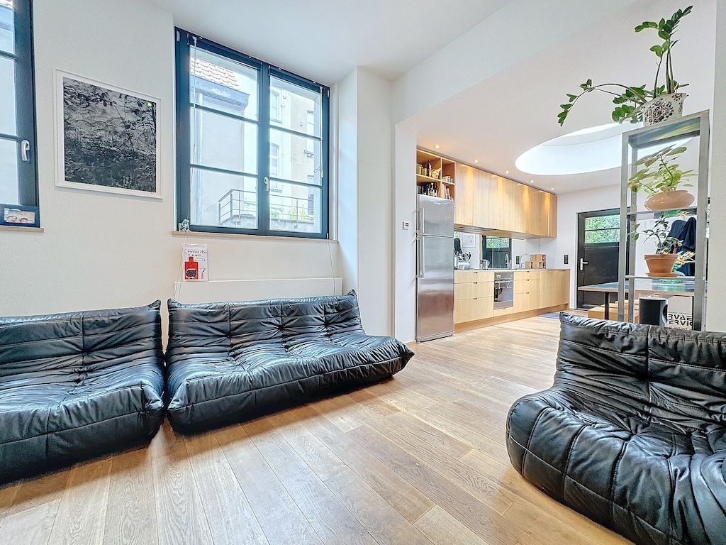 Salon contemporain lumineux avec canapés noirs, cuisine en bois clair, plantes, grande fenêtre industrielle.