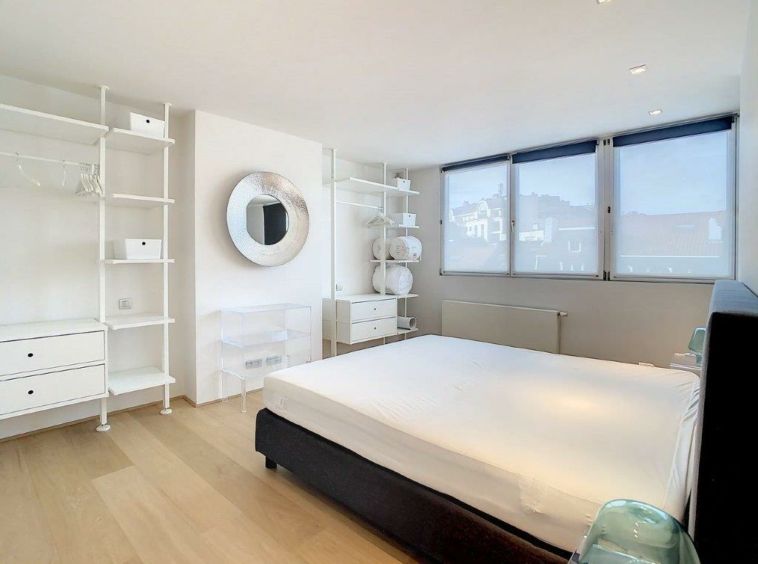 Chambre minimaliste lumineuse avec lit nu, étagères blanches, miroir rond et vue naturelle.