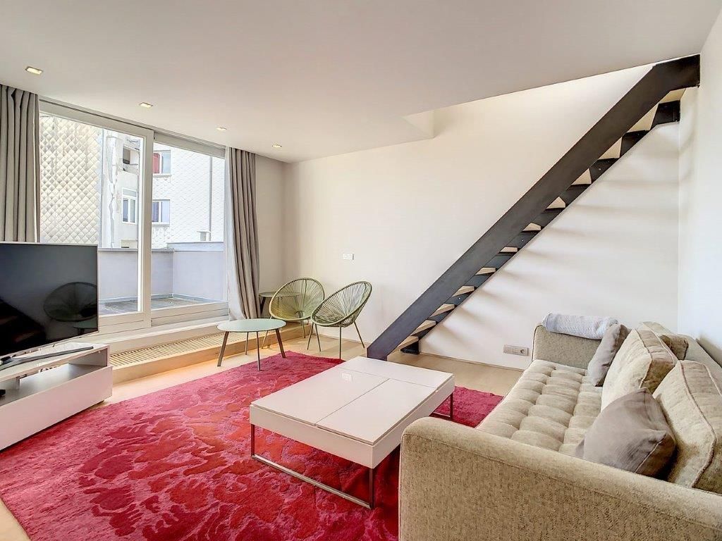 Salon moderne lumineux avec canapé beige, tapis rouge, fauteuils verts et escalier en bois dark.