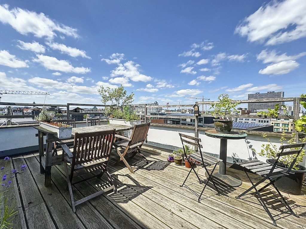 Terrasse urbaine ensoleillée avec mobilier rustique, plantes en pots et vue sur la ville.