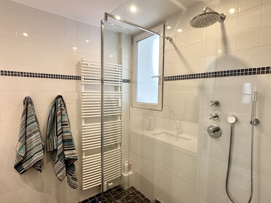 Salle de douche moderne avec carrelage blanc, douche à pluie, radiateur et serviettes colorées.