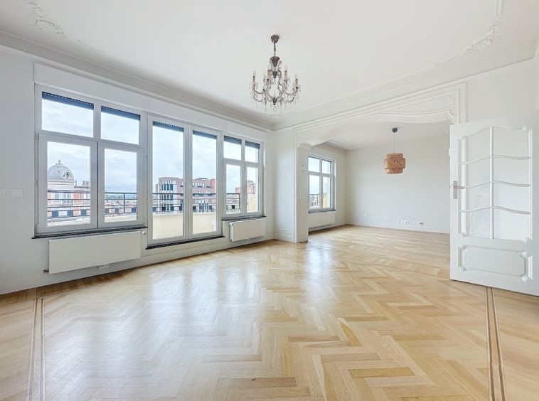 Salle lumineuse avec parquet chevron, large fenêtres, lustre élégant et moulures classiques.