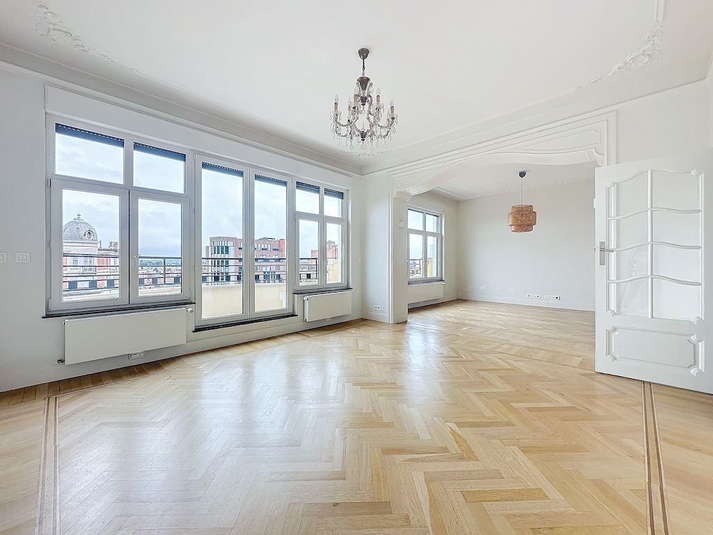 Salle lumineuse avec parquet chevron, large fenêtres, lustre élégant et moulures classiques.