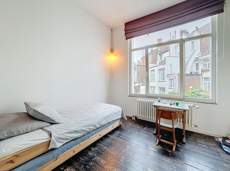 Chambre minimaliste lumineuse avec vue urbaine, lit en bois clair, stores violets et ambiance chaleureuse.