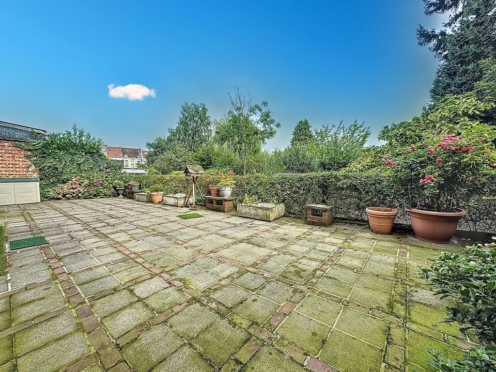 Patio spacieux entouré de plantes fleuries, sous un ciel bleu lumineux et arboré, parfait pour se détendre.