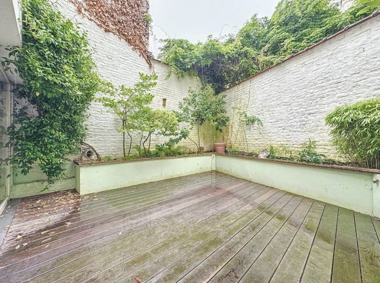 Cour de détente rustique avec murs blancs, végétation luxuriante et terrasse en bois humide.