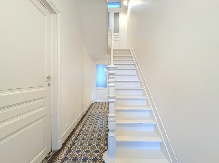 Hall lumineux avec escalier blanc, murs neutres et carrelage décoratif bleu et blanc.