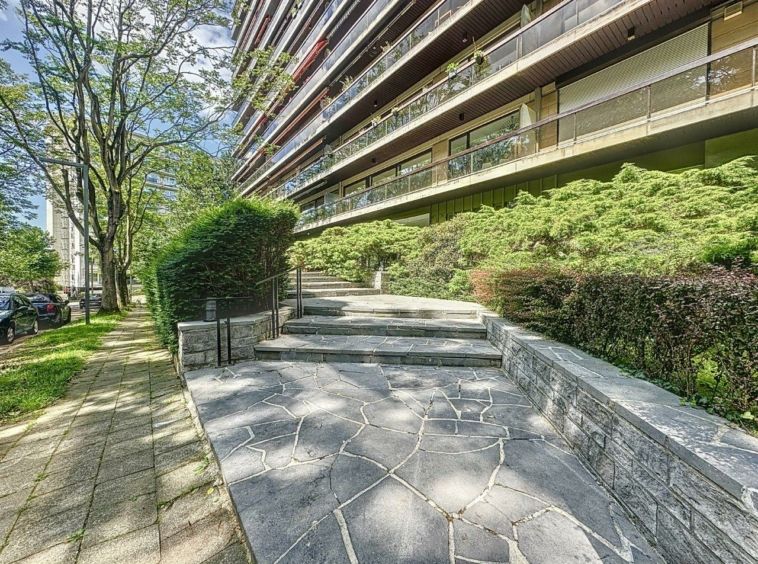 Résidence moderne avec balcons, façade en verre, jardin urbain et promenade arborée.