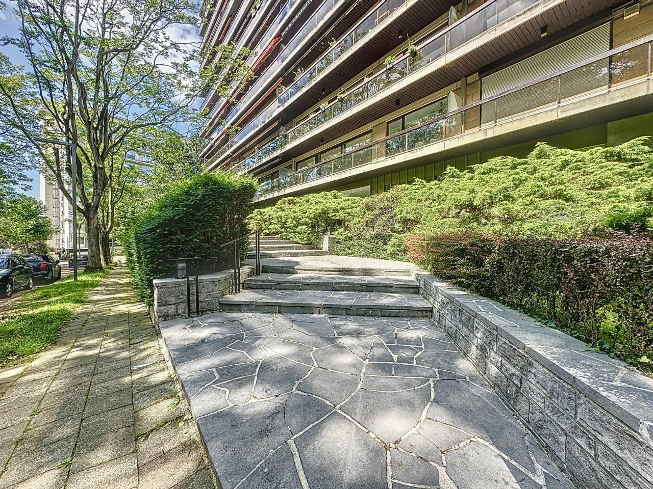 Résidence moderne avec balcons, façade en verre, jardin urbain et promenade arborée.