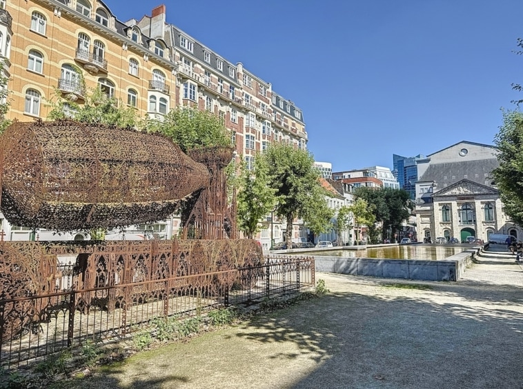 Parc urbain ensoleillé avec sculpture métallique, étang, arbres et résidence moderne.