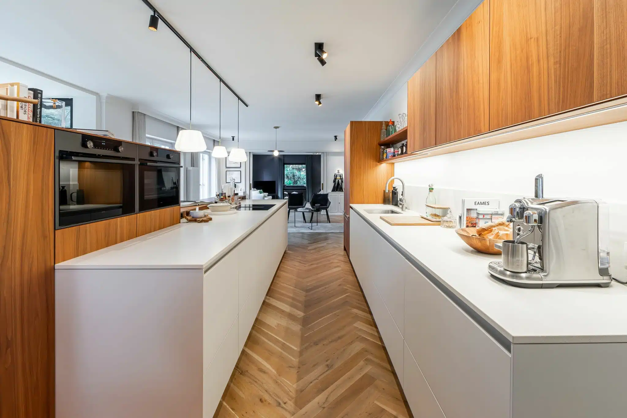 Cuisine moderne chaleureuse avec plan blanc, meubles en bois, éclairage suspendu et parquet à chevrons.