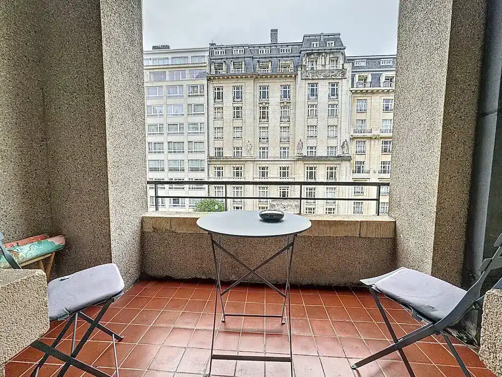 Balcon urbain cosy avec vue architecturale, mobilier minimaliste, sous ciel gris, étage moderne et historique.