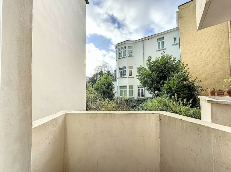 Balcon urbain avec vue sur immeuble, jardin vert, ciel bleu et nuages blancs.