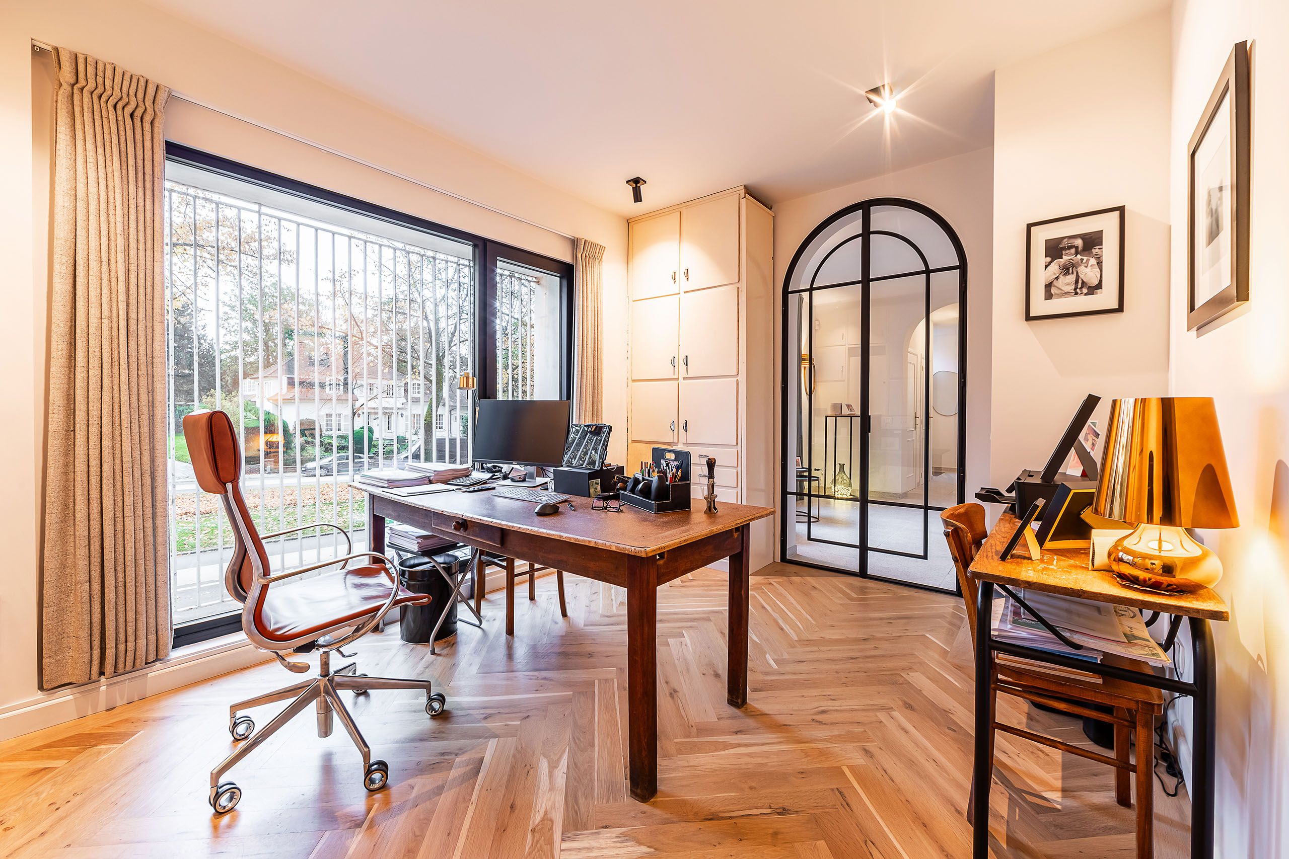 Espace de travail moderne avec vue sur jardin, bureau en bois, chaise en cuir rouge, et décoration élégante.