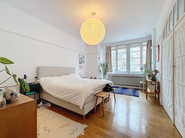 Chambre lumineuse élégante avec lit central, plantes vertes, tapis coloré et mobilier en bois.