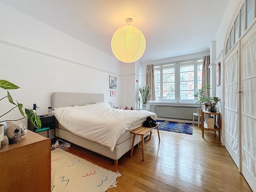 Chambre lumineuse élégante avec lit central, plantes vertes, tapis coloré et mobilier en bois.