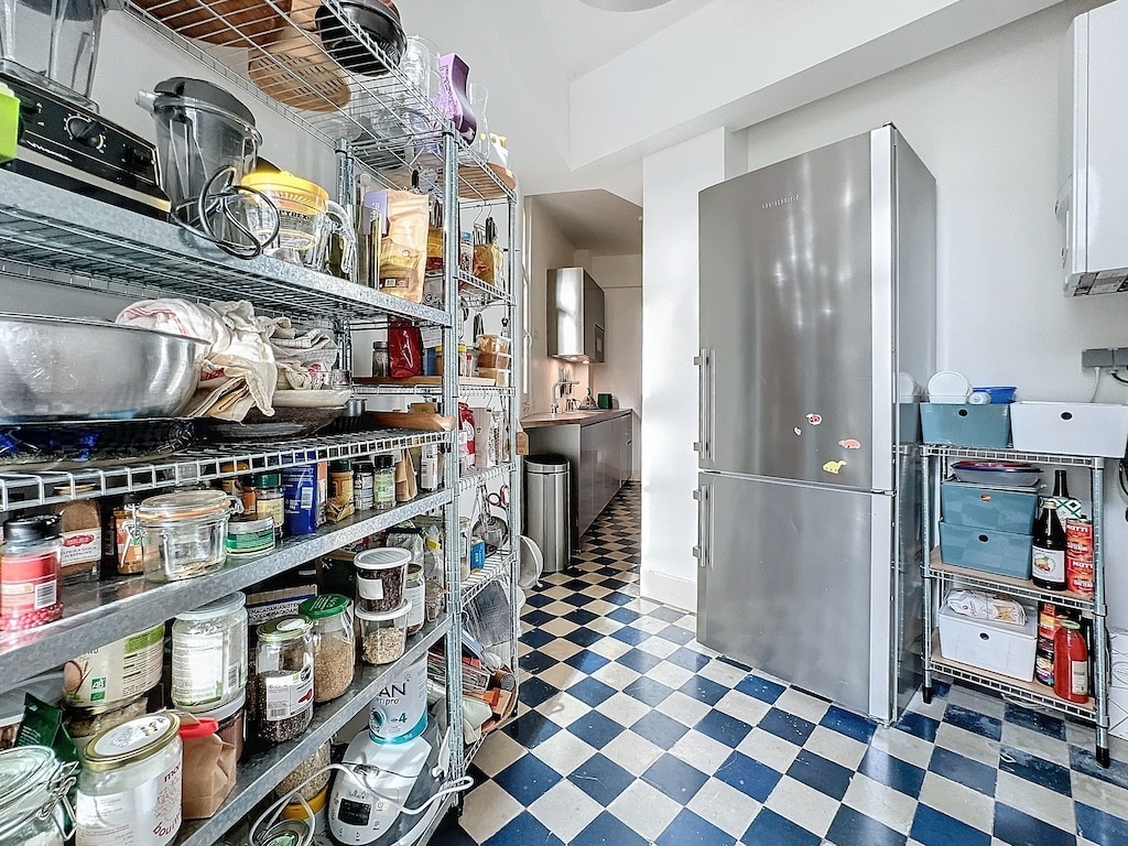 Cuisine moderne avec sol en carreaux bleu et blanc, rangement métal, réfrigérateur inox, lumière naturelle.