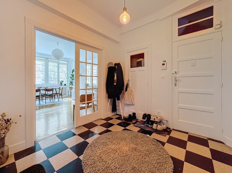 Entrée lumineuse et élégante avec carreaux noirs et blancs, plantes, porte-manteaux et porte en verre dépoli.