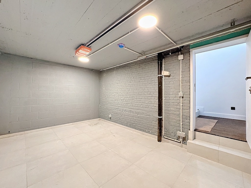 Salle de sous-sol moderne, lumineuse, polyvalente, avec sol clair, murs gris et tuyaux visibles.