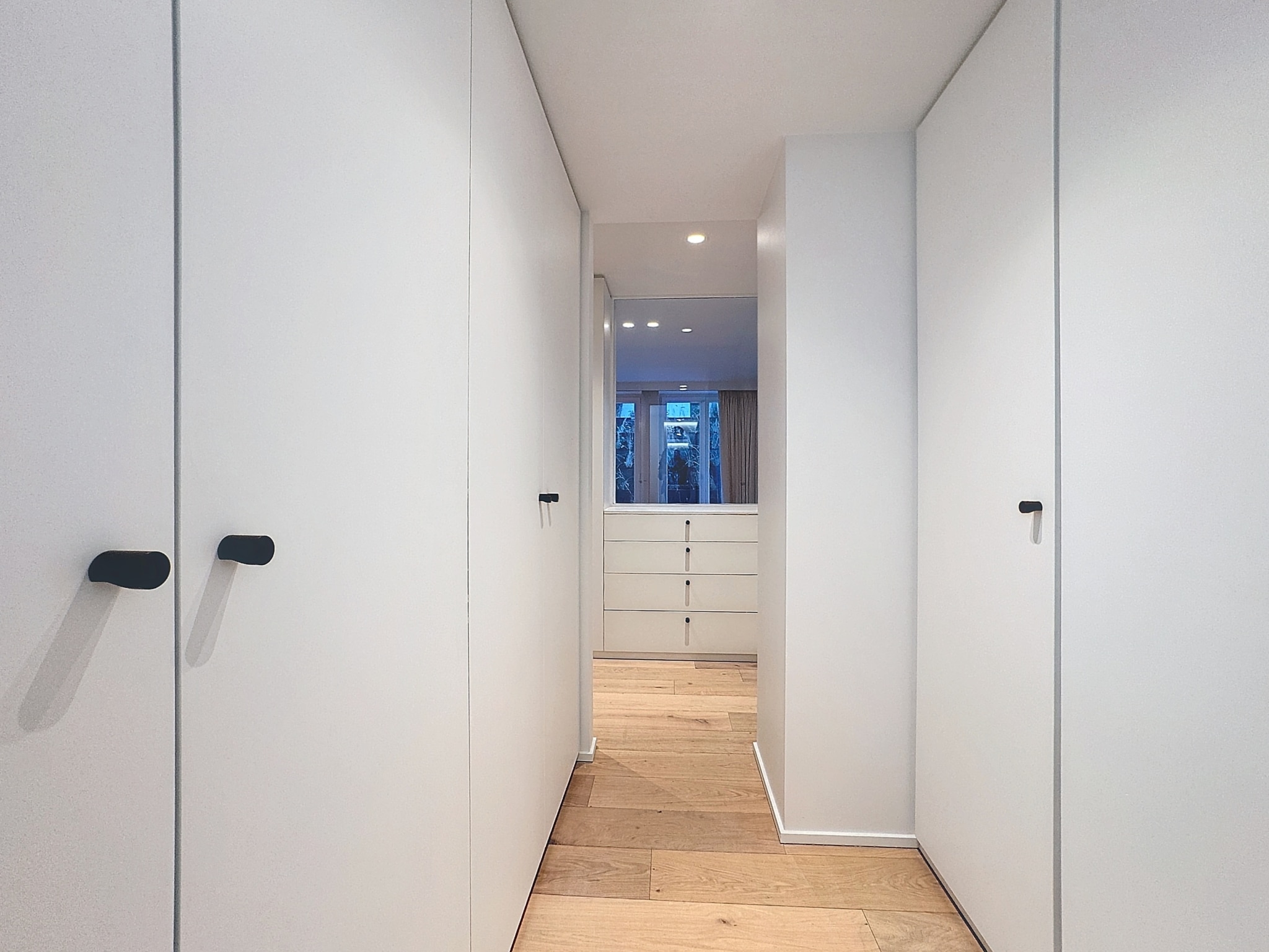 Hall moderne minimaliste en blanc, bois clair, lumière naturelle et poignées noires élégantes.