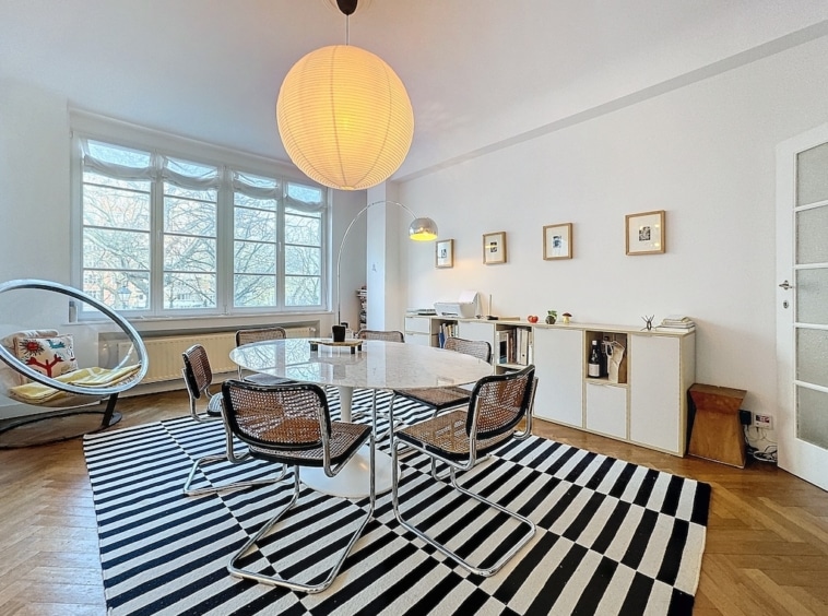 Salon lumineux et moderne avec table ronde, chaises élégantes, tapis noir et blanc, ambiance chaleureuse.