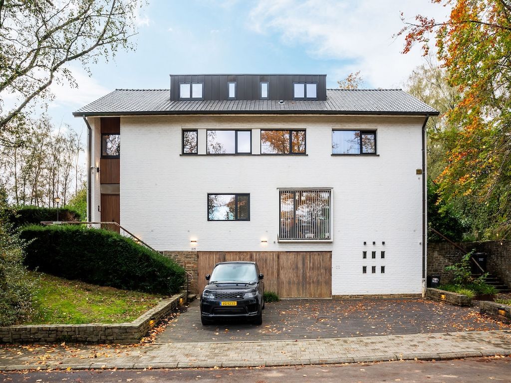Maison moderne avec façade blanche, grandes fenêtres, jardin automnal et voiture noire élégante.