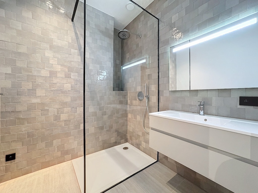 Salle de bain moderne avec douche à l’italienne, murs gris, meuble blanc et éclairage LED.