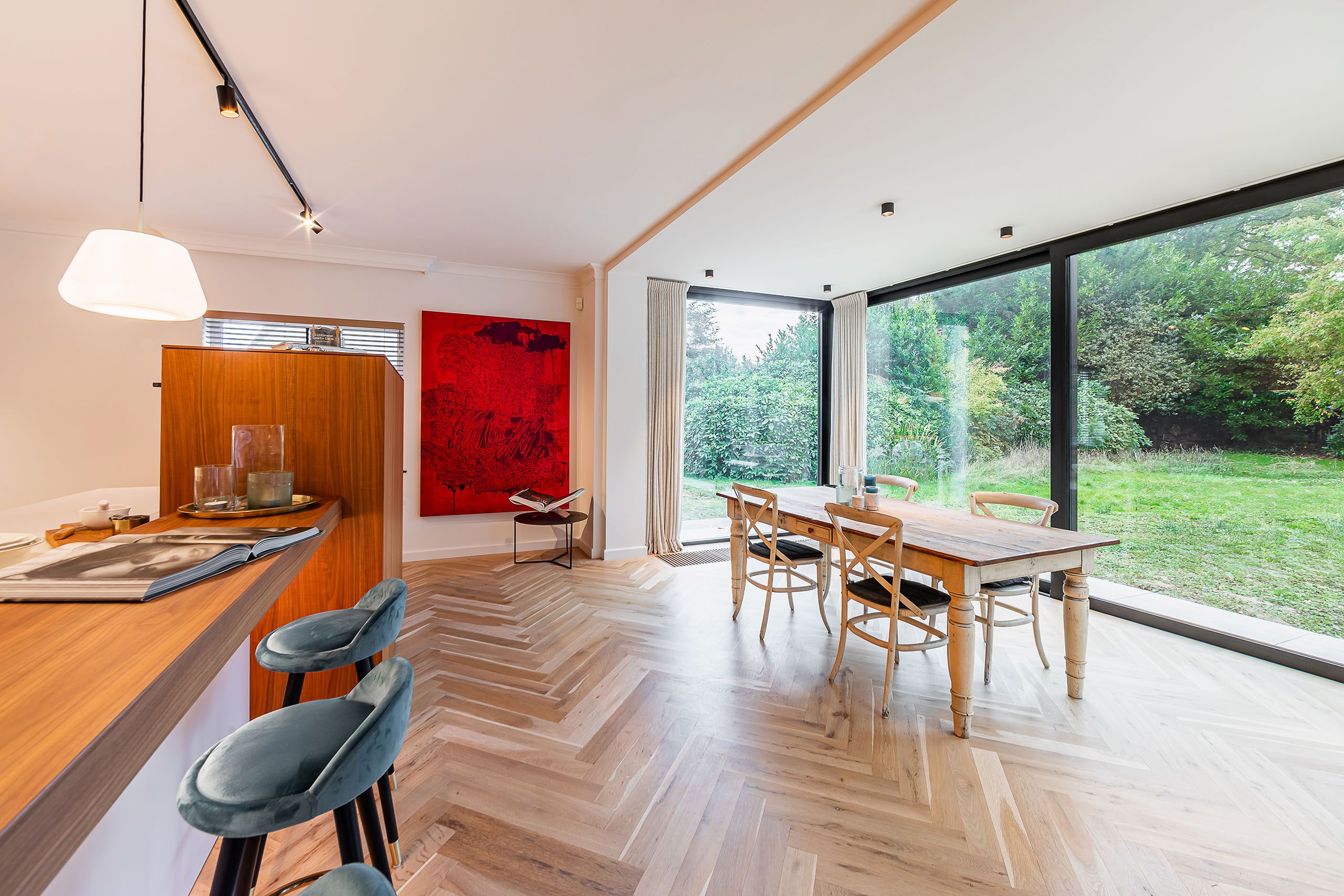 Salle à manger moderne avec vue sur jardin, mobilier en bois, accents bleus et œuvre colorée.