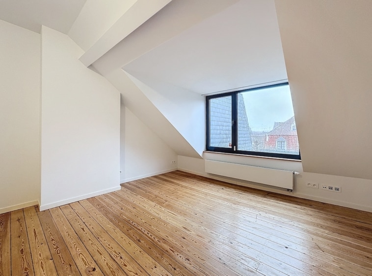 Attique lumineux avec grande fenêtre, sol en parquet chaleureux, espace polyvalent et vue urbaine.