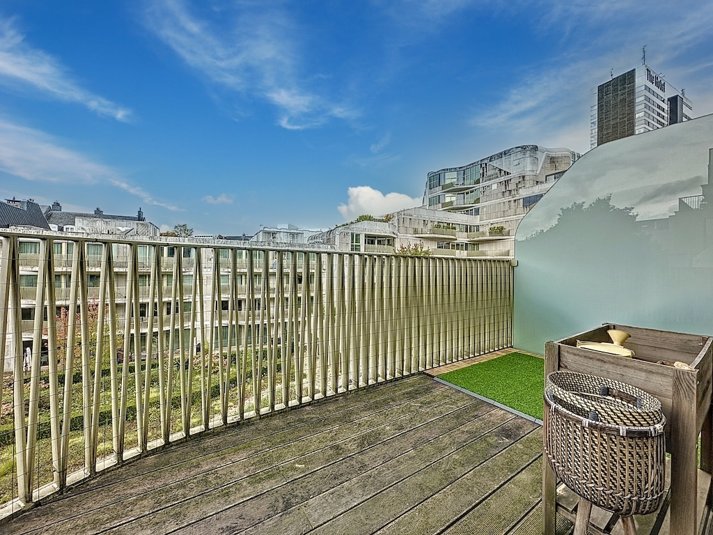 Balcon urbain avec sol en bois, espace vert et vue sur ville moderne sous ciel bleu.