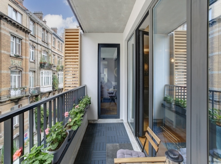 Balcon moderne avec plantes, fauteuil, vue urbaine, mélange d’architecture contemporaine et classique.