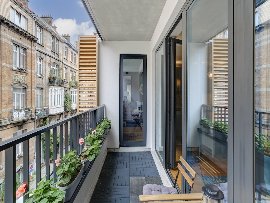 Balcon moderne avec plantes, fauteuil, vue urbaine, mélange d’architecture contemporaine et classique.