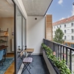 Balcon moderne avec plantes, fauteuil bleu, vue urbaine, lumière naturelle, ambiance zen et conviviale.